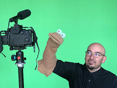 Man holding a soft puppet that covers his arm performing in front of a green screen. There is a camera he is performing to.